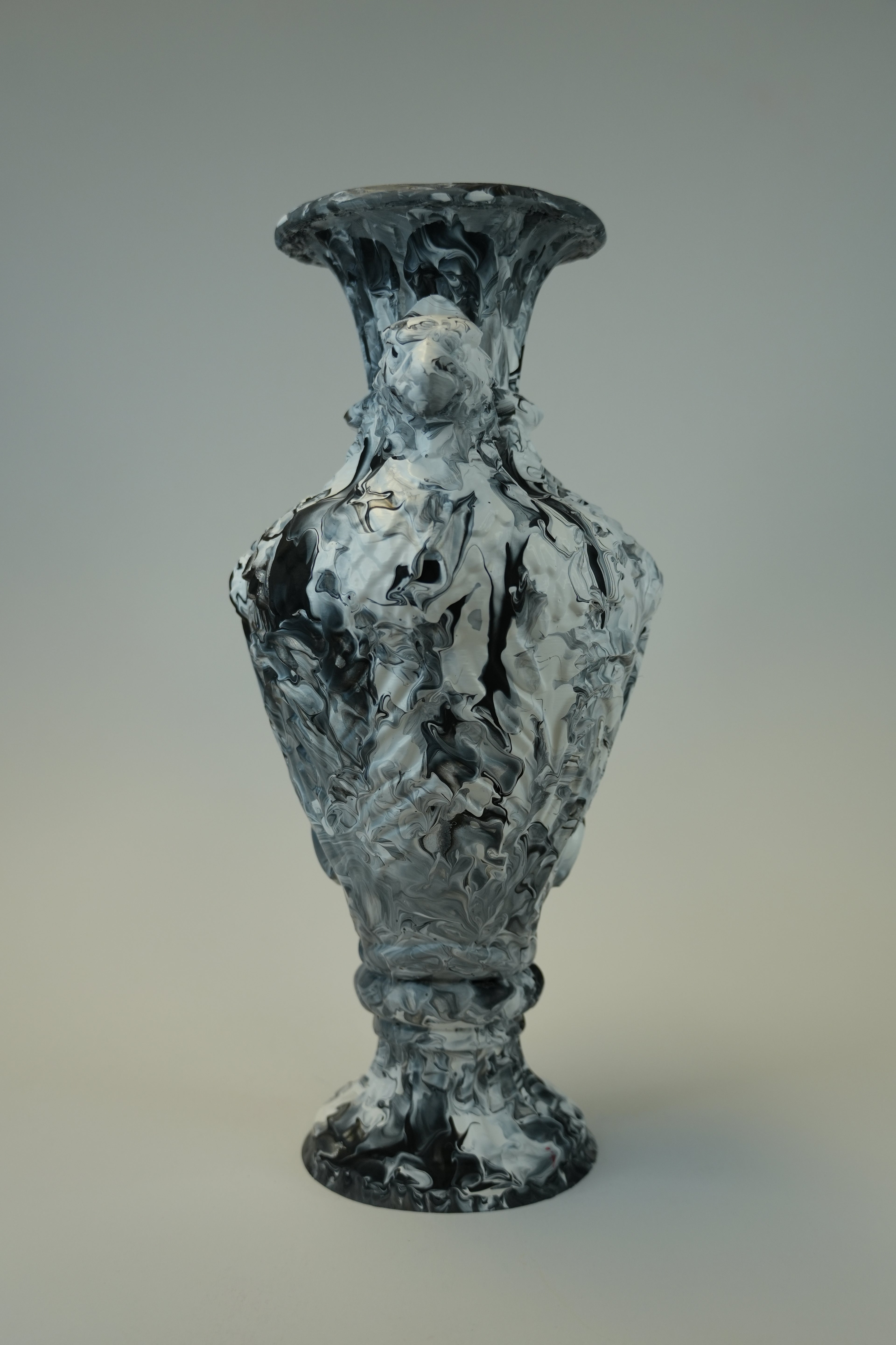 A sculptural vase with a flared rim and pedestal base, featuring an abstract marbled pattern in black, white, and gray, photographed against a neutral gray gradient background.