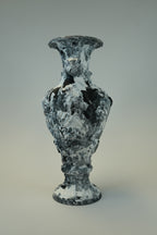 A sculptural vase with a textured, marbled appearance in black, white, and gray, set against a neutral light gray background.