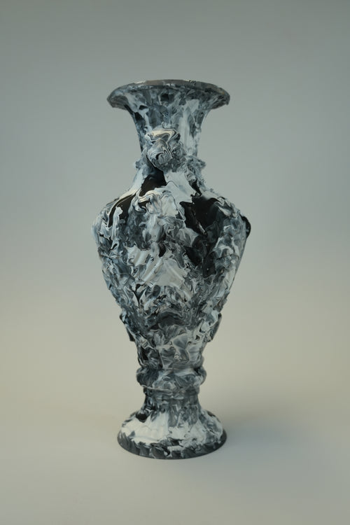 A sculptural vase with a textured, marbled appearance in black, white, and gray, set against a neutral light gray background.