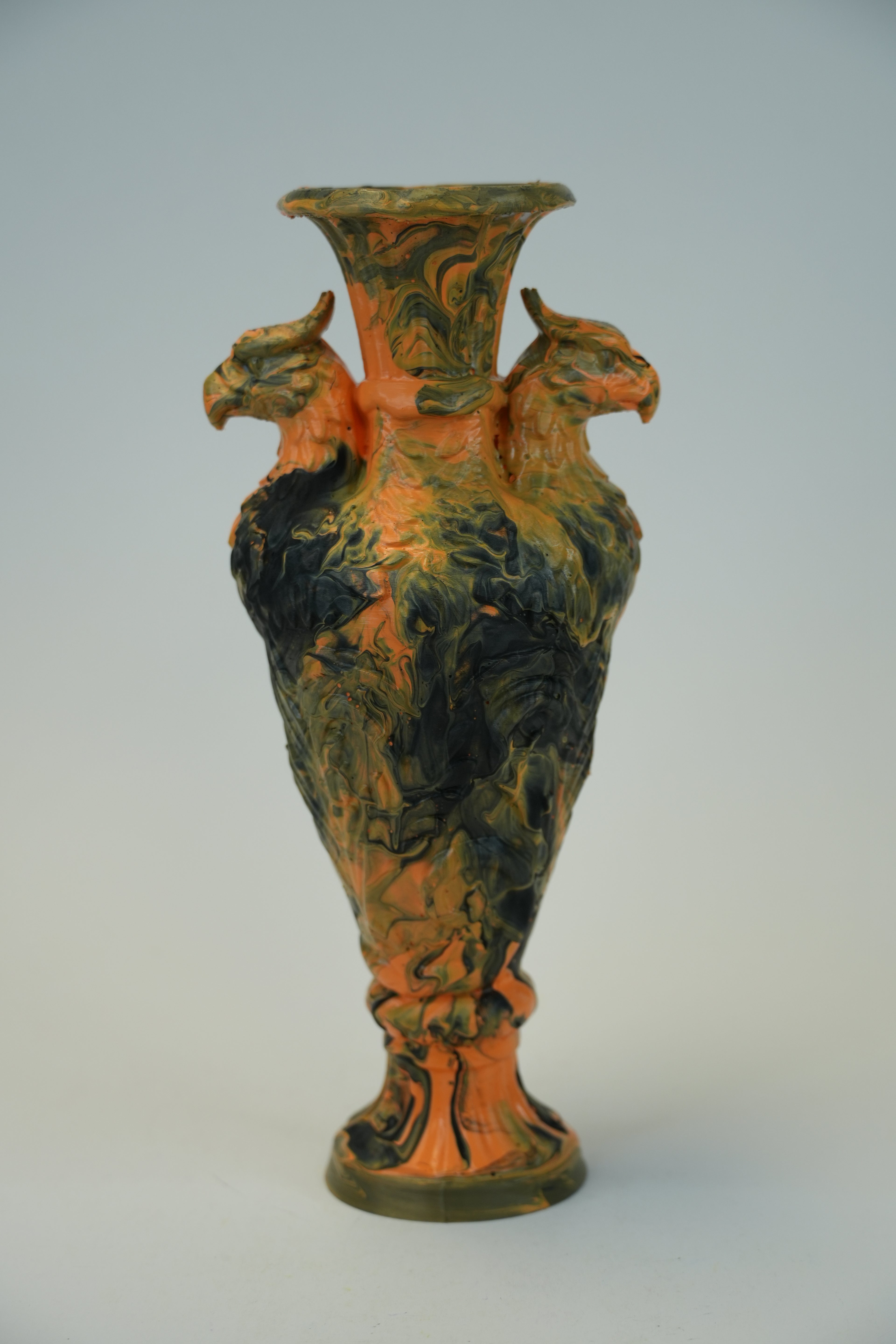 A decorative ceramic vase with a flared rim and a sculpted body featuring two rooster heads on the sides, covered in a marbled glaze of orange, black, and olive green, set against a plain light gray background.