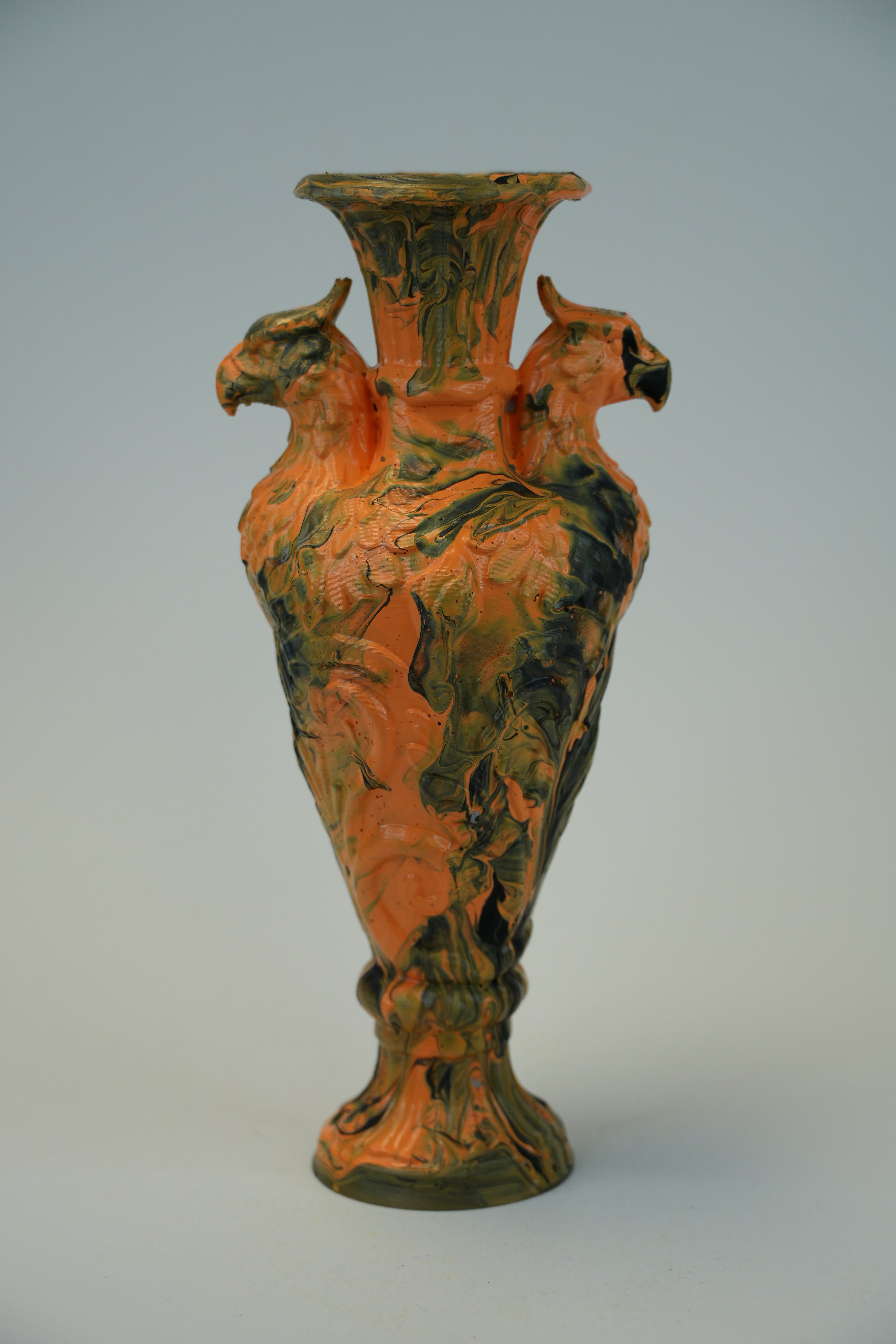 A decorative ceramic vase with a flared rim, a narrow neck, and two bird figures serving as handles, decorated with a marbled pattern of orange, green, and dark brown glaze, set against a neutral light gray background.