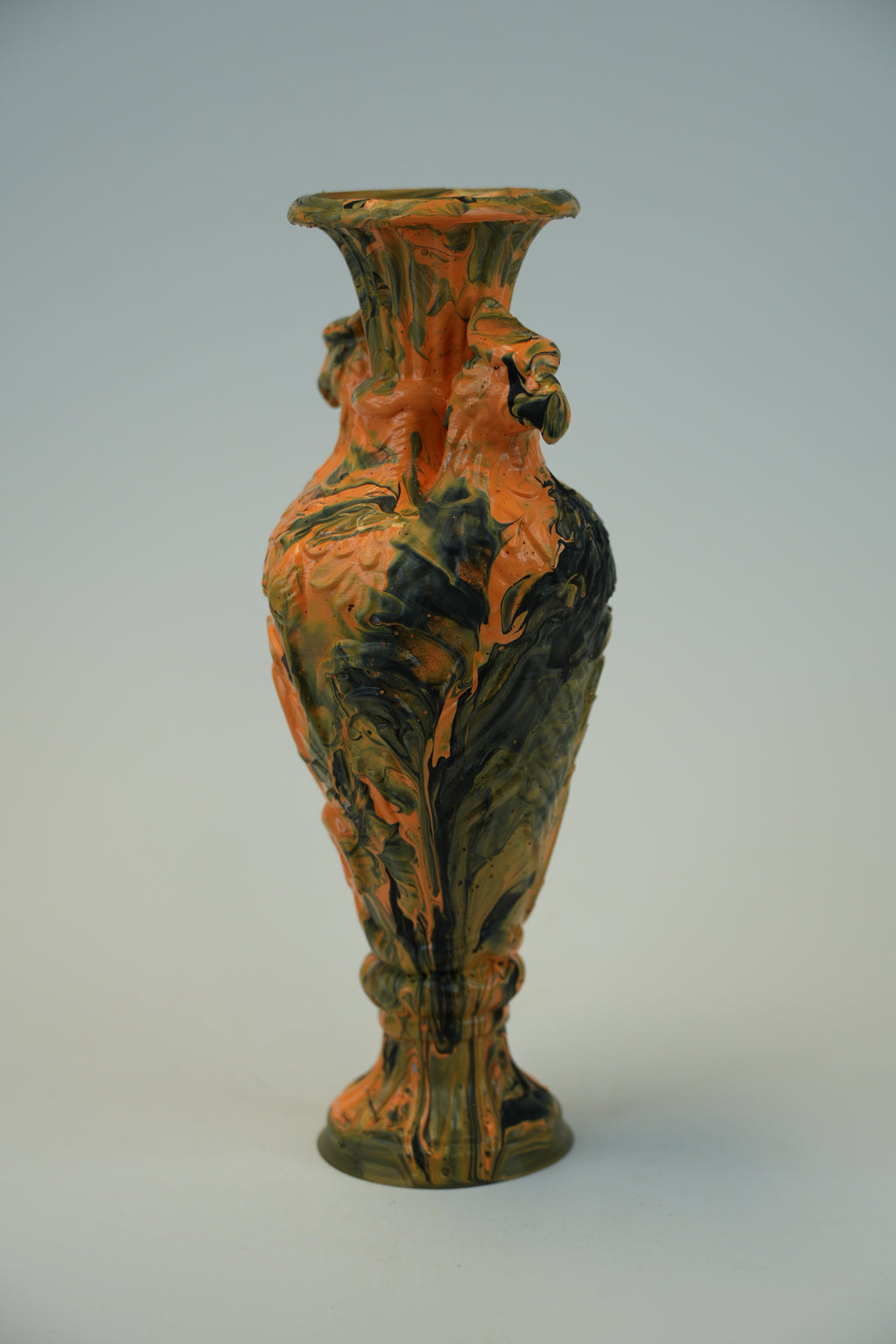 An ornate ceramic vase with a flared rim and sculpted decorative details, featuring a marbled glaze in orange, black, and brown tones, set against a neutral light gray background.