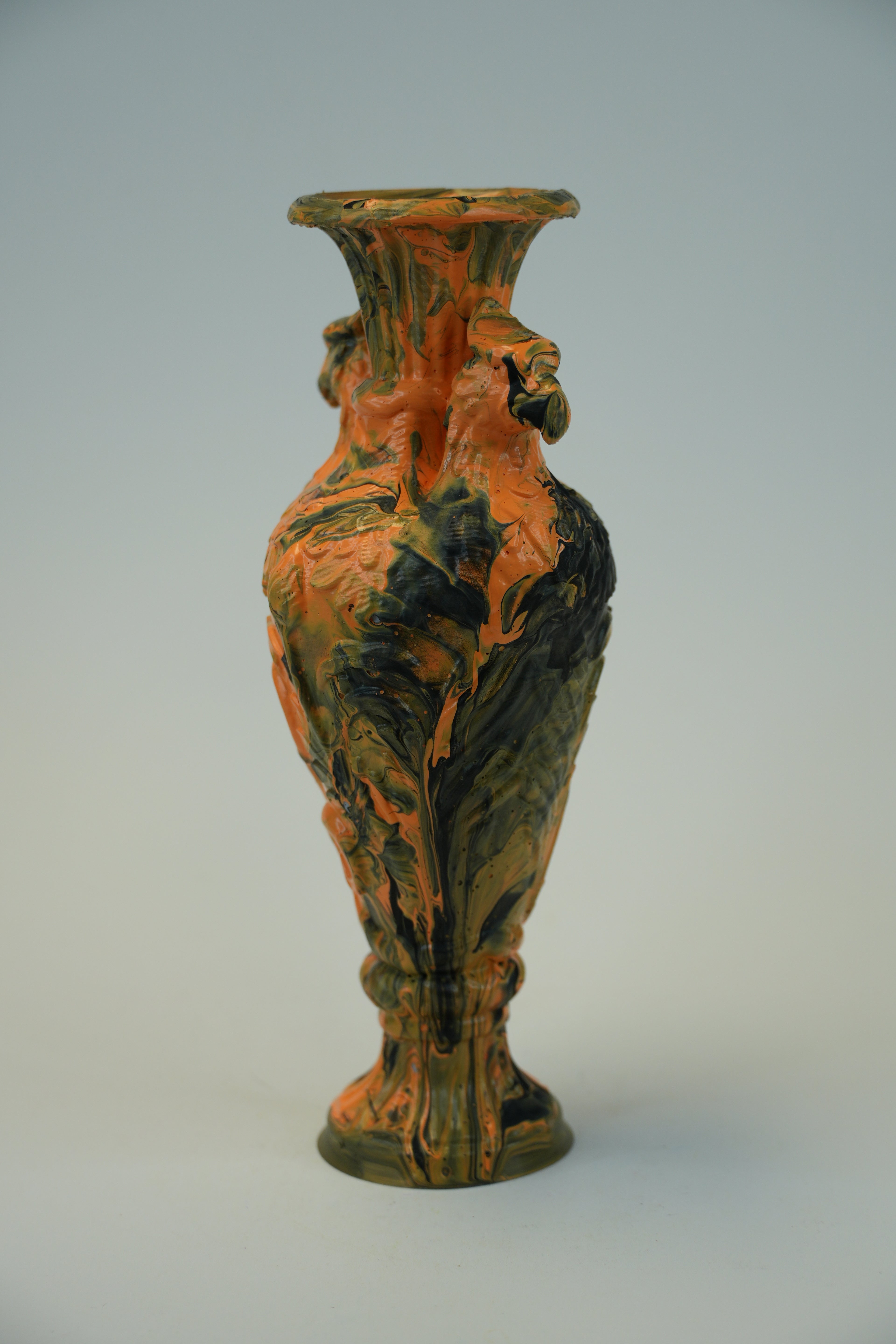 An ornate ceramic vase with a flared rim and sculpted decorative details, featuring a marbled glaze in orange, black, and brown tones, set against a neutral light gray background.
