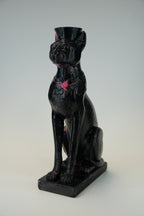 A glossy black cat figurine wearing a small top hat and a pink bow tie, seated on a rectangular base against a light gray studio background.