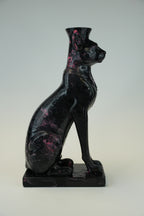A glossy black cat figurine with pink marbled inclusions, shown in a seated pose on a matching rectangular base against a neutral light gray background.