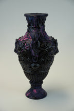 An ornate dark purple and black glass vase with embossed floral relief patterns, set against a neutral light gray background.