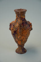 An ornate vintage-style glass vase with a bulbous body, flared neck, and pedestal base, decorated with raised floral and scroll motifs. The vase has a warm amber-brown tint with darker reddish-brown shading, photographed against a plain light gray studio background.