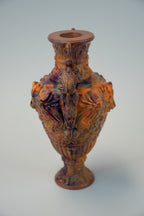 An ornate ceramic vase with a flared rim and a sculpted, highly textured body featuring organic, marbled patterns in terracotta orange, brown, and deep purple, set against a neutral light gray background.