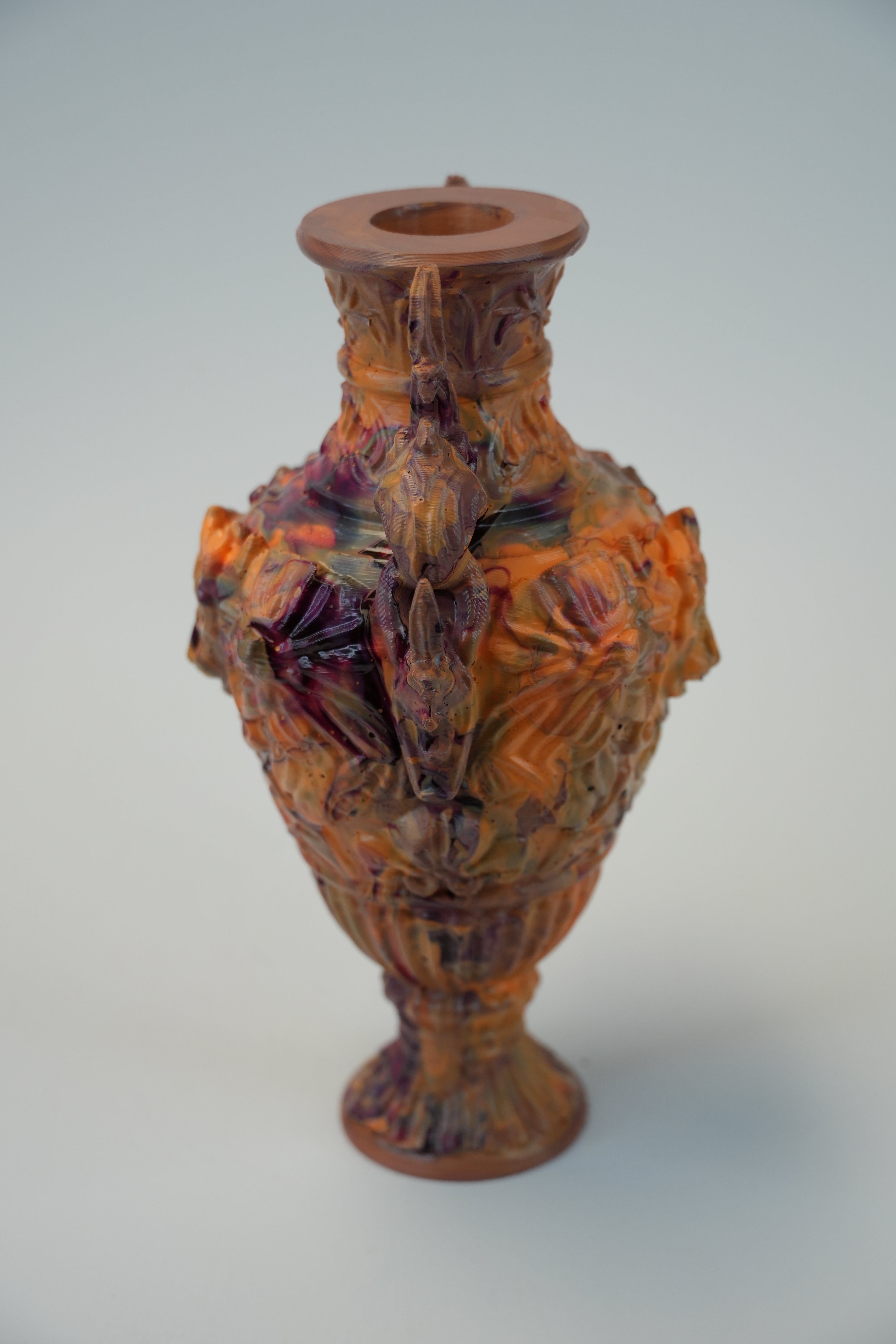 An ornate ceramic vase with a flared rim and a sculpted, highly textured body featuring organic, marbled patterns in terracotta orange, brown, and deep purple, set against a neutral light gray background.