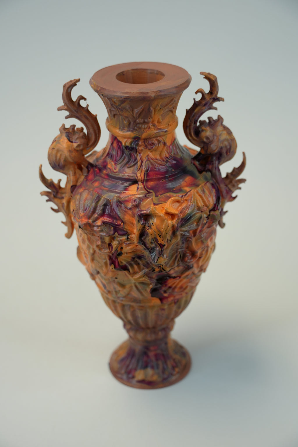 An ornate sculptural vase with a flared rim, featuring intricate relief carvings of dragons or mythical creatures in warm brown, orange, and red tones against a plain light background.