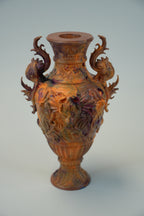 An ornate carved ceramic vase with two dragon-like handles, featuring a glossy finish with iridescent copper, gold, and purple tones, set against a plain light gray background.