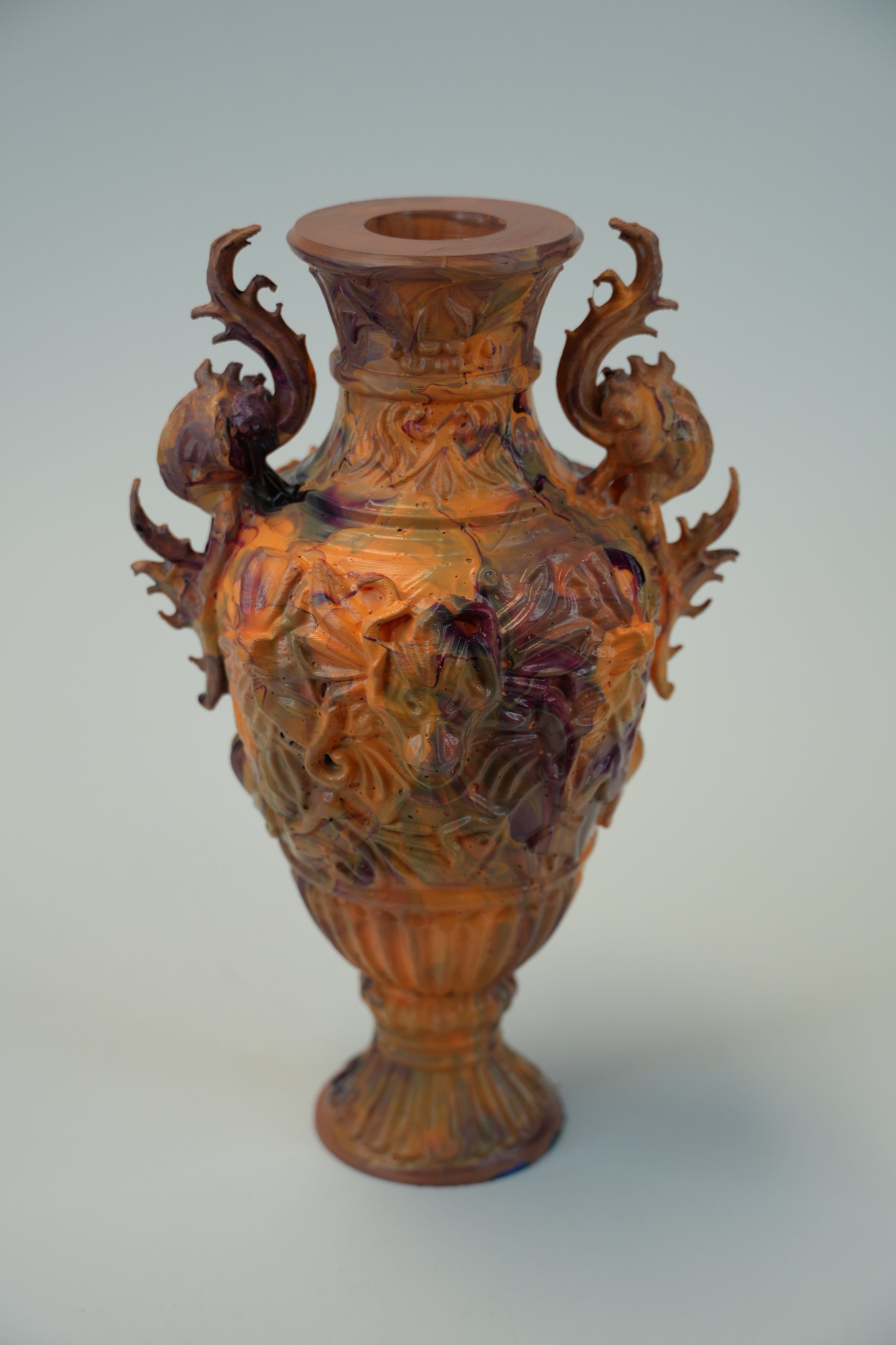 An ornate carved ceramic vase with two dragon-like handles, featuring a glossy finish with iridescent copper, gold, and purple tones, set against a plain light gray background.