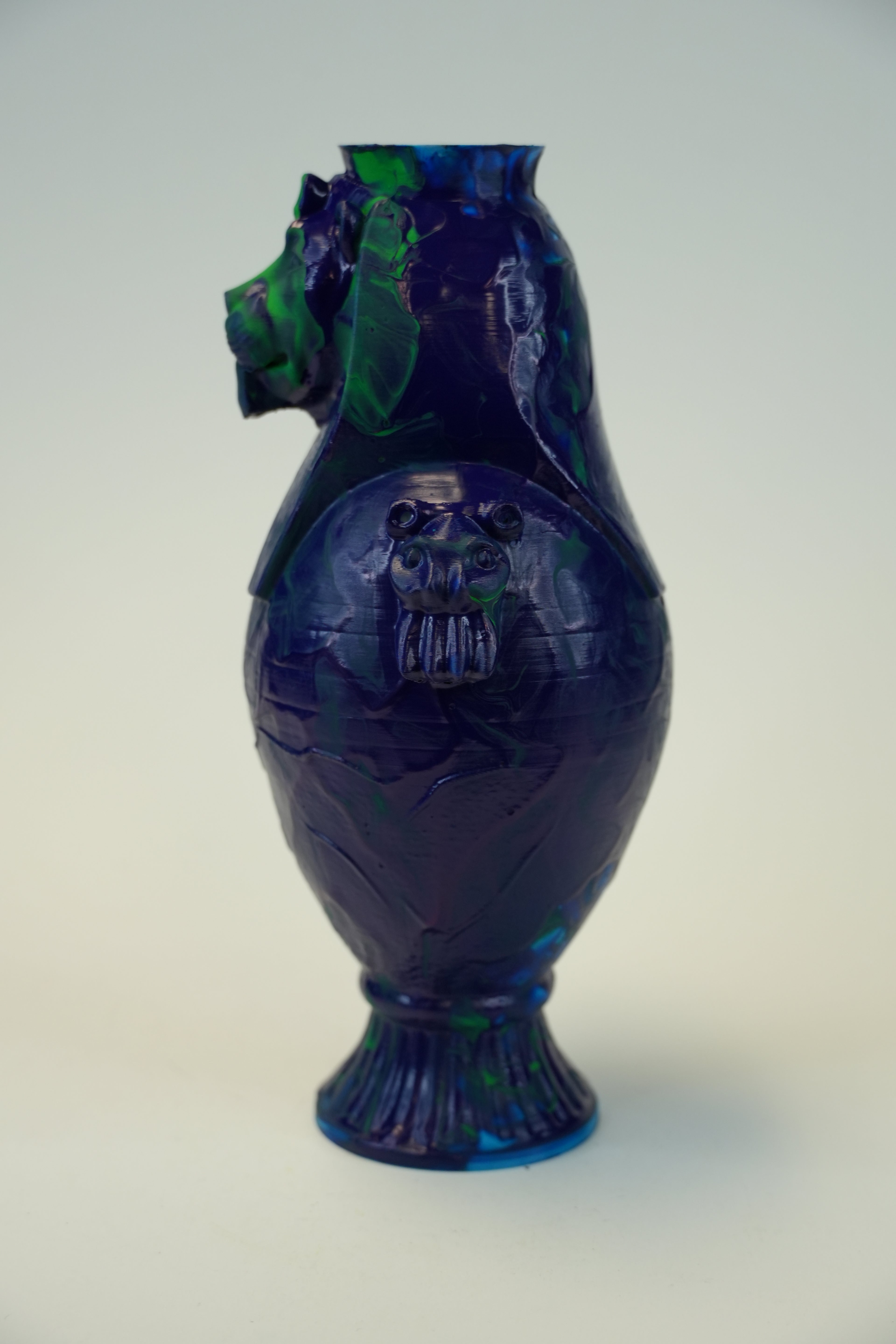 A decorative ceramic vase with a deep cobalt blue and green marbled glaze, featuring a sculpted lion head spout and a matching lion face relief on the front, set against a plain light background.