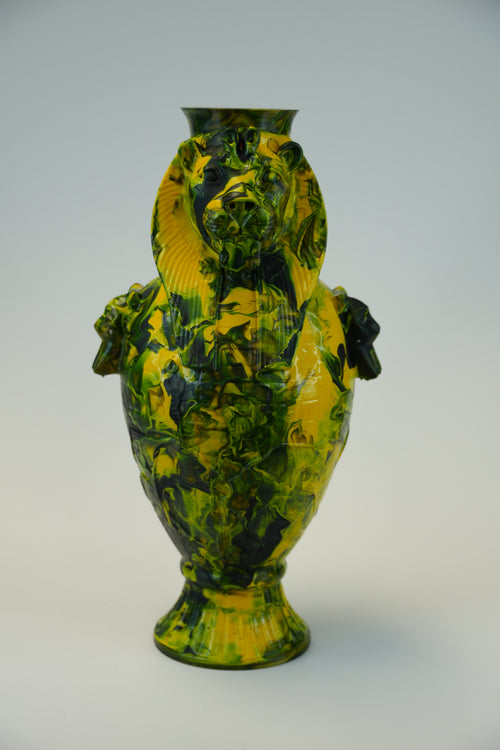 A decorative ceramic vase with a glossy yellow and dark green marbled pattern, featuring sculpted lion heads on the sides and a flared base, photographed against a neutral light gray background.