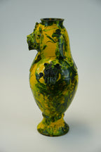 A vintage-style ceramic vase with a glossy marbled yellow and dark green glaze, featuring a sculpted lion head spout and a black floral relief on the front, set against a neutral light gray background.