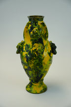 A decorative ceramic vase with sculpted lion heads on the shoulders and a flared pedestal base, decorated in a marbled pattern of yellow, green, and dark brown/black.