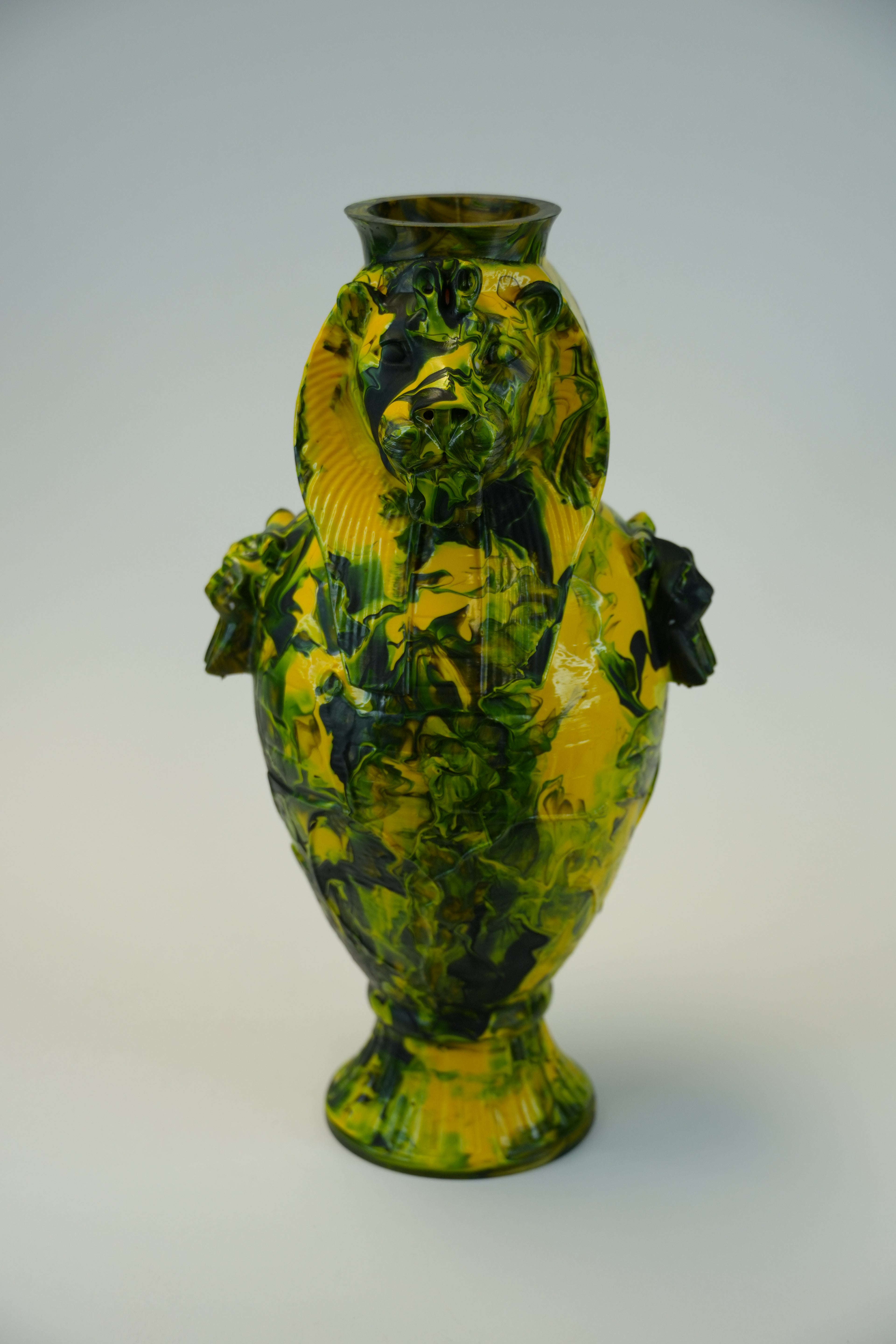 A decorative ceramic vase with sculpted lion heads on the shoulders and a flared pedestal base, decorated in a marbled pattern of yellow, green, and dark brown/black.
