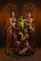 Three women in ornate gold and brown Egyptian-inspired costumes pose with a green and gold decorative vase against a dark brown backdrop with large lion head graphics.