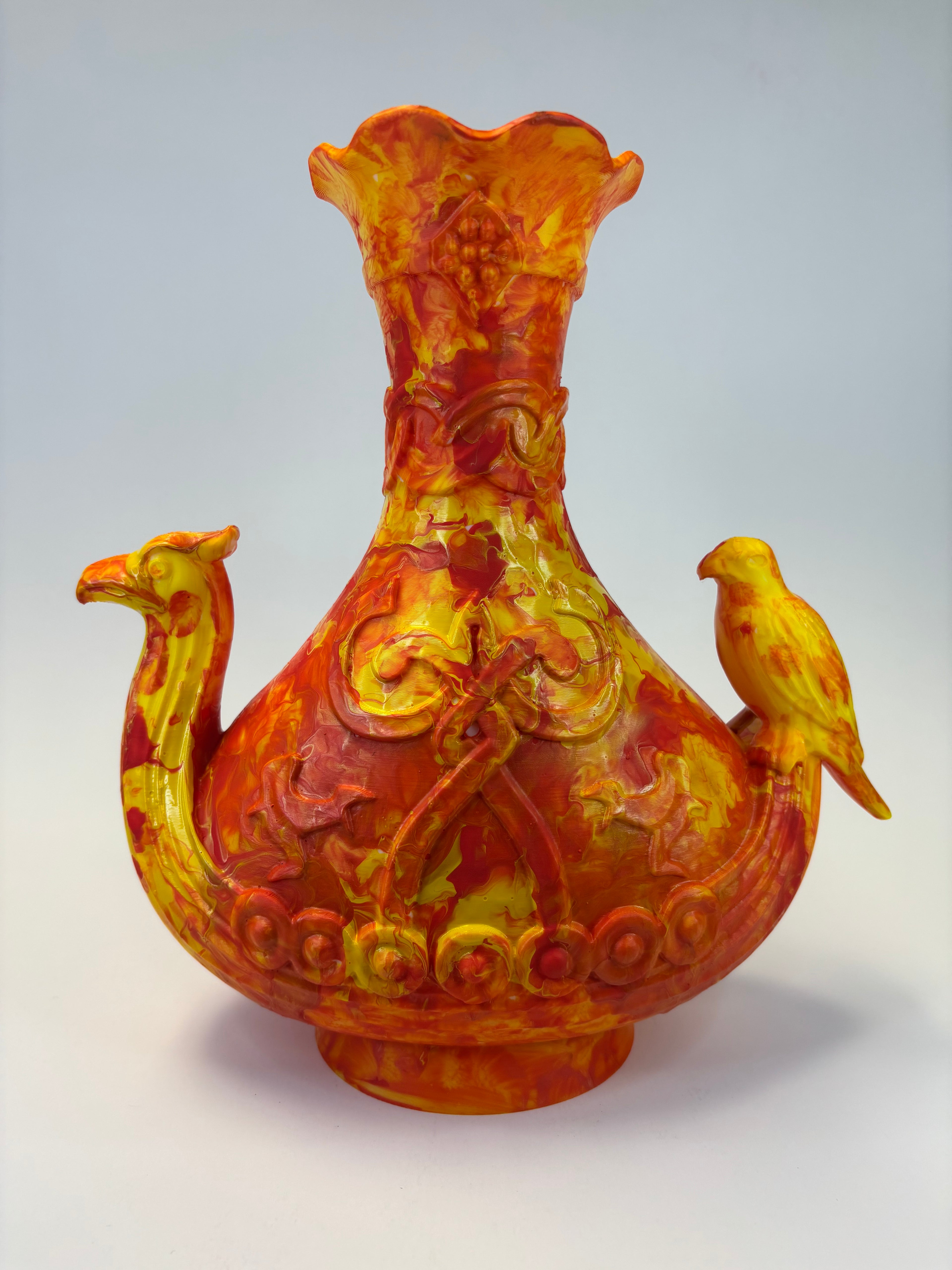A sculptural ceramic vase with a bulbous body, flared rim, and two bird-shaped handles, painted in a marbled mix of yellow, orange, and red glaze against a plain light background.