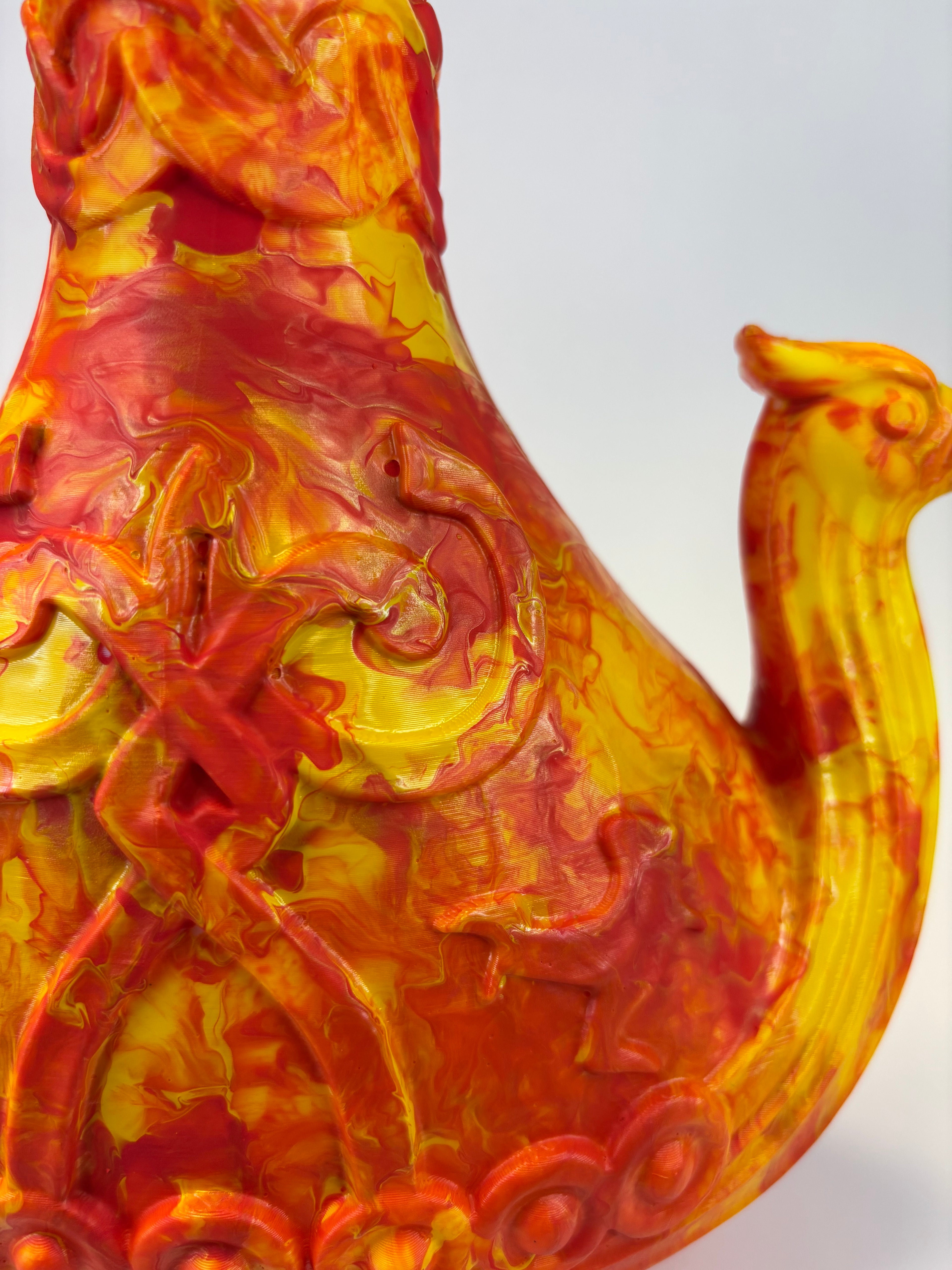 A close-up of a sculptural ceramic vase or pitcher with a camel-like handle, decorated in swirling red, orange, and yellow glaze patterns against a light gray background.