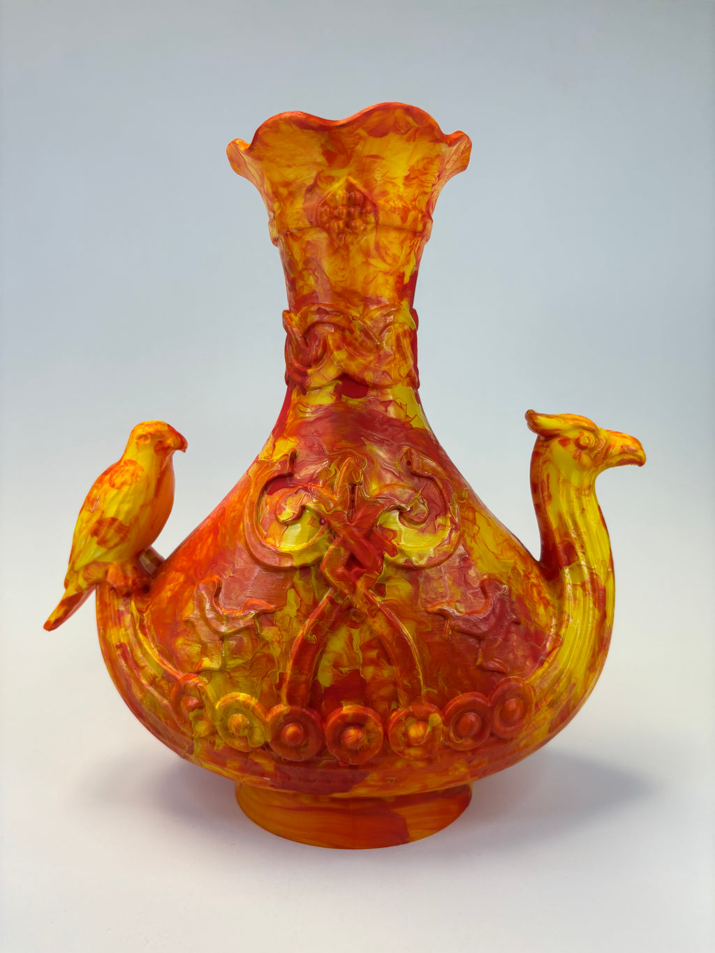 An ornate sculptural vase in marbled yellow and orange glass, featuring a flared neck, a rounded body with embossed scrollwork and circular motifs, and two bird figures perched on the sides, photographed against a light gray studio background.