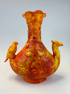 An ornate sculptural vase in marbled yellow and orange glass, featuring a flared neck, a rounded body with embossed scrollwork and circular motifs, and two bird figures perched on the sides, photographed against a light gray studio background.