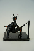 A small black fantasy figurine of a horned demon-like character crouching and holding a whip, mounted on a rectangular base against a plain light gray background.