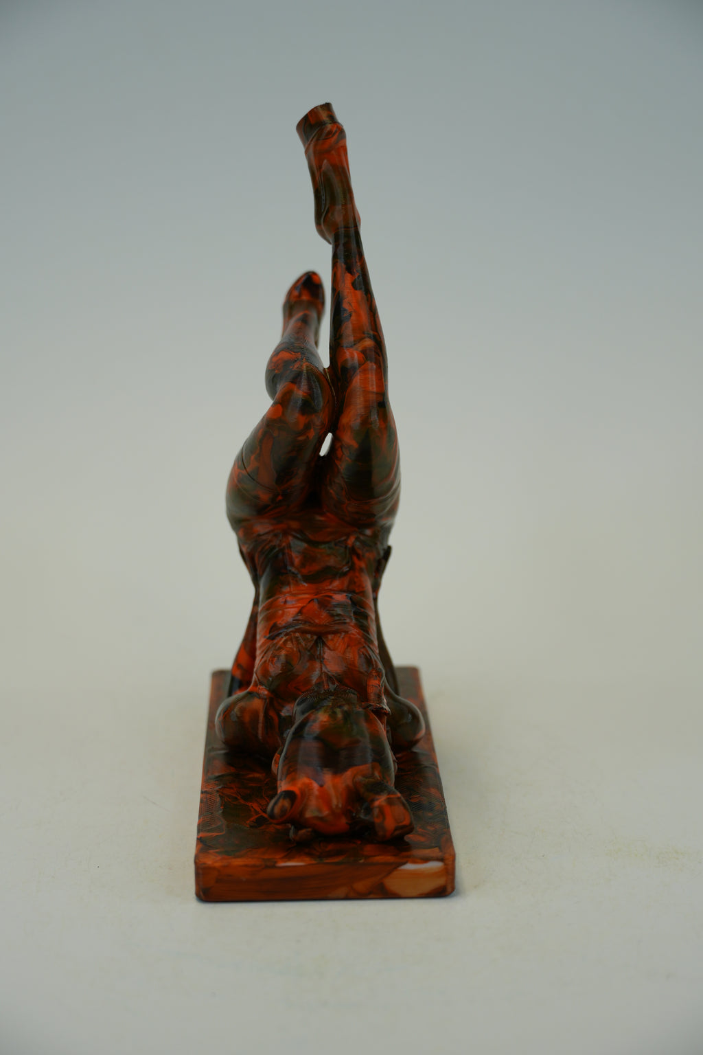 A small sculptural figurine of a human figure in a dynamic pose, made from a glossy marbled resin or stone with swirling orange, red, and dark brown tones, standing on a rectangular base against a plain light gray background.
