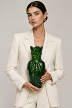 A woman in a cream-colored blazer holds a glossy, sculptural green glass vase shaped like a beetle against a neutral light gray background.