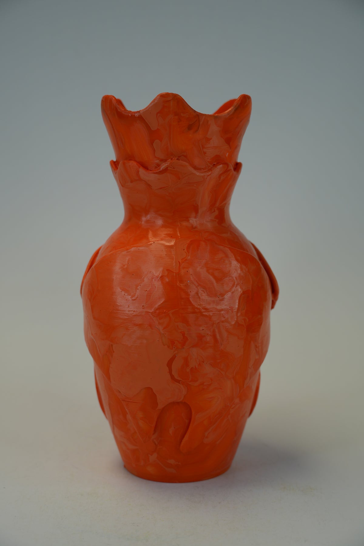 An orange-red ceramic vase with a textured, organic surface and a flared, scalloped rim, photographed against a neutral gray studio background.