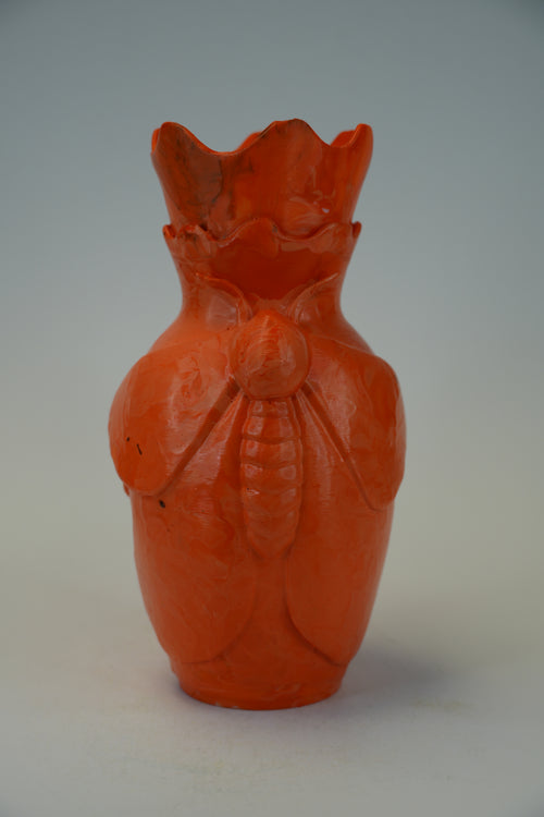 An orange-red carved ceramic vase shaped like a beetle, photographed against a neutral gray background.