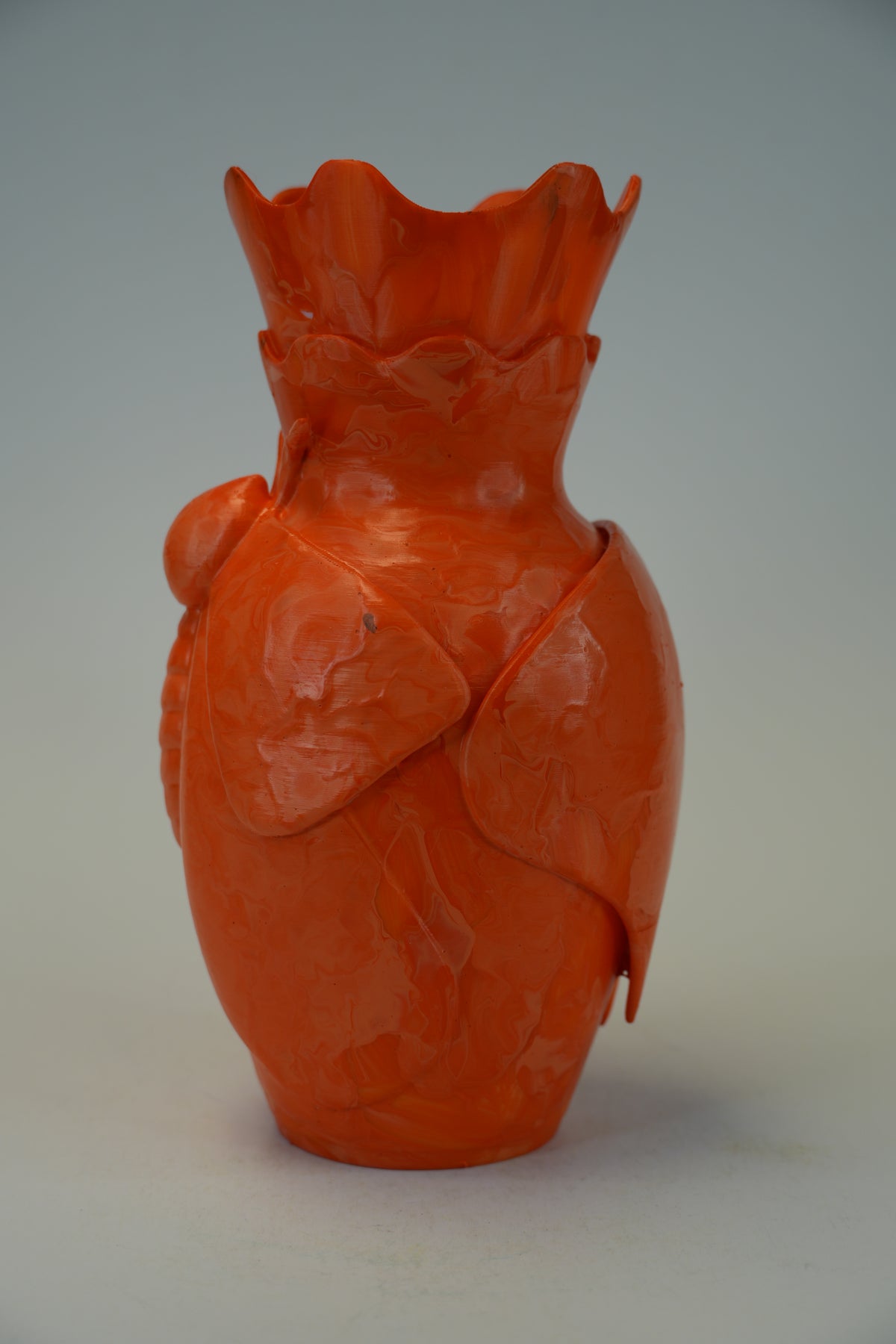 An orange sculptural ceramic vase with a textured, organic surface and a flared, ruffled rim, photographed against a neutral gray background.