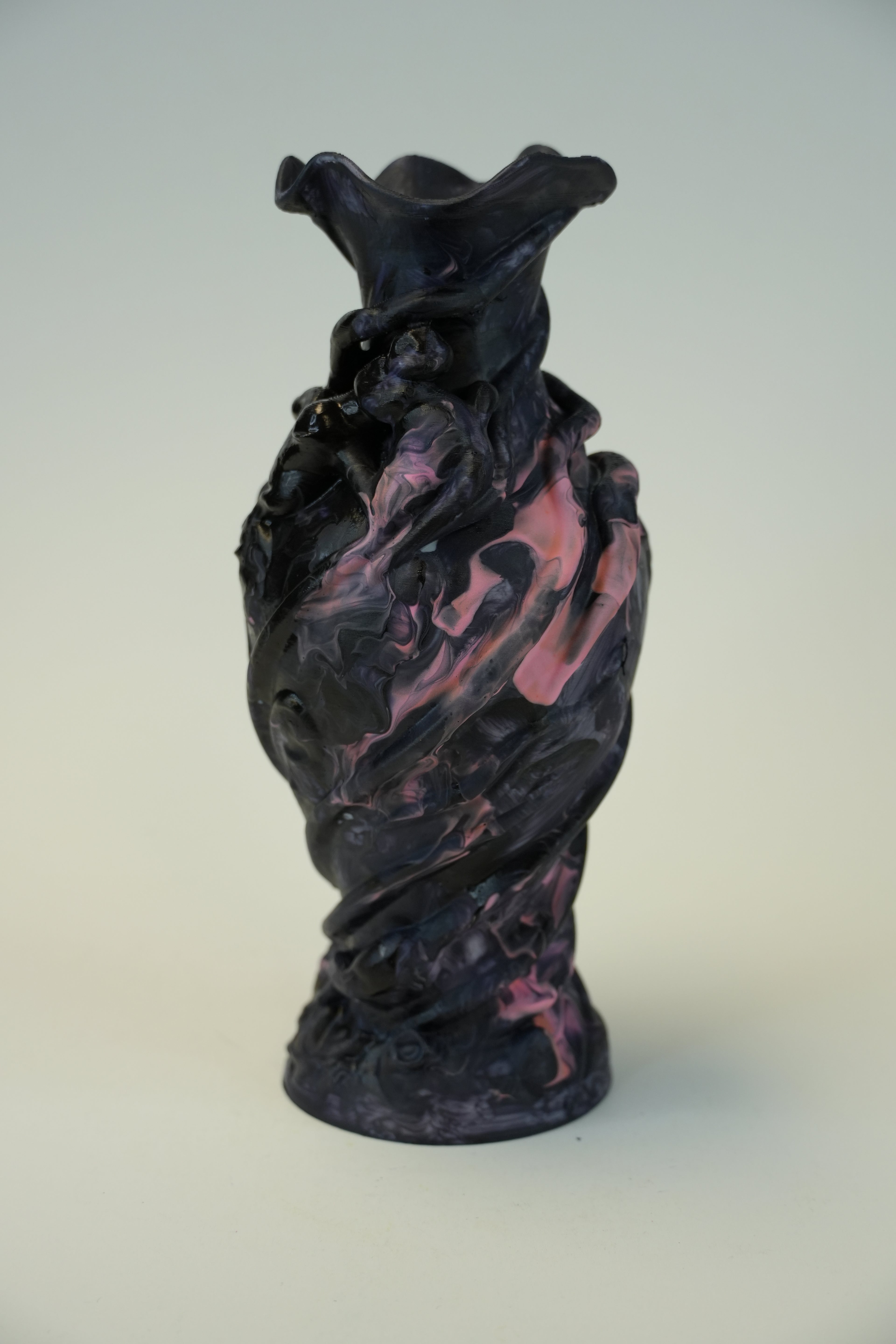 A sculptural vase with a dark, marbled texture in black and deep purple, accented with pink swirls, standing on a light neutral background.