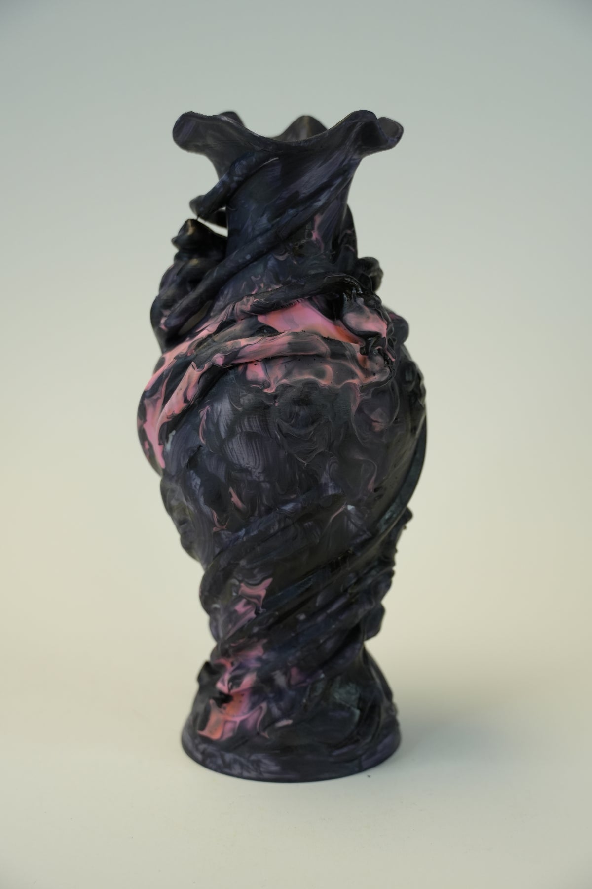 An abstract sculptural vase with a dark charcoal-black base and swirling pink and purple marbled accents, photographed against a neutral light gray background.