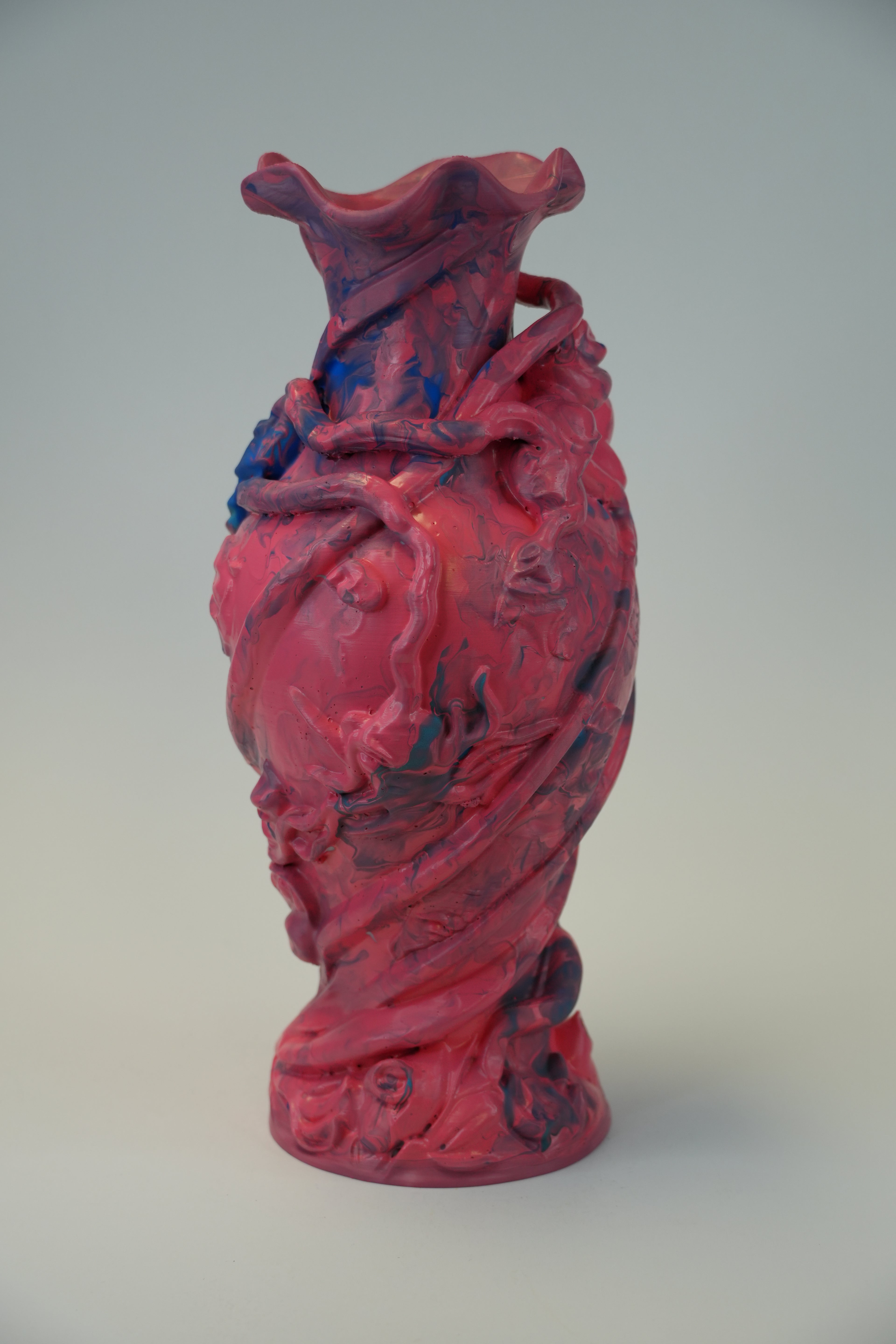 A sculptural ceramic vase with a flared rim, featuring a highly textured, organic surface with swirling patterns of magenta pink, red, and dark blue, set against a neutral light gray background.