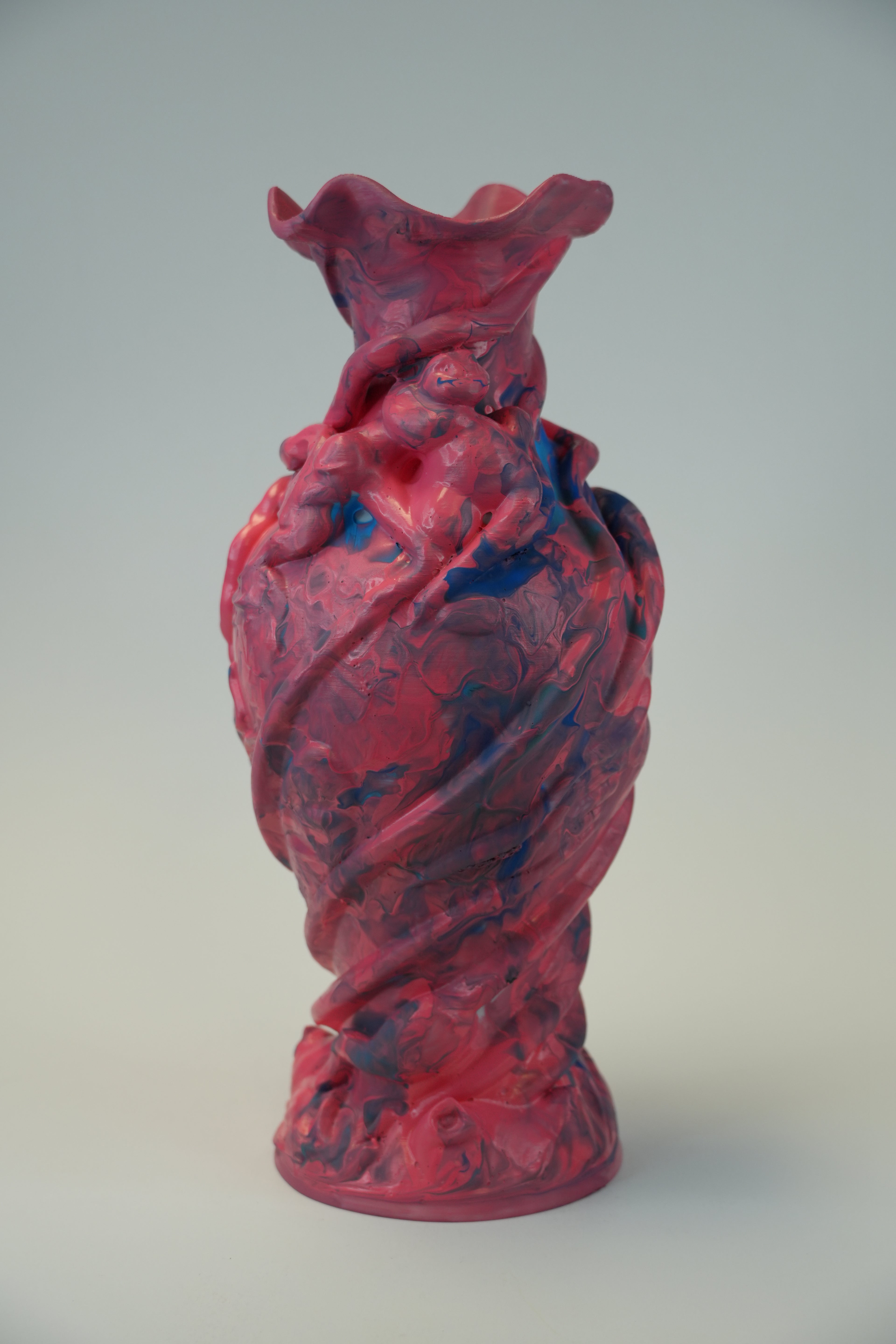 A sculptural art piece featuring a twisted, organic form with a marbled pattern of red, pink, and blue paint, mounted on a plain light gray wall.