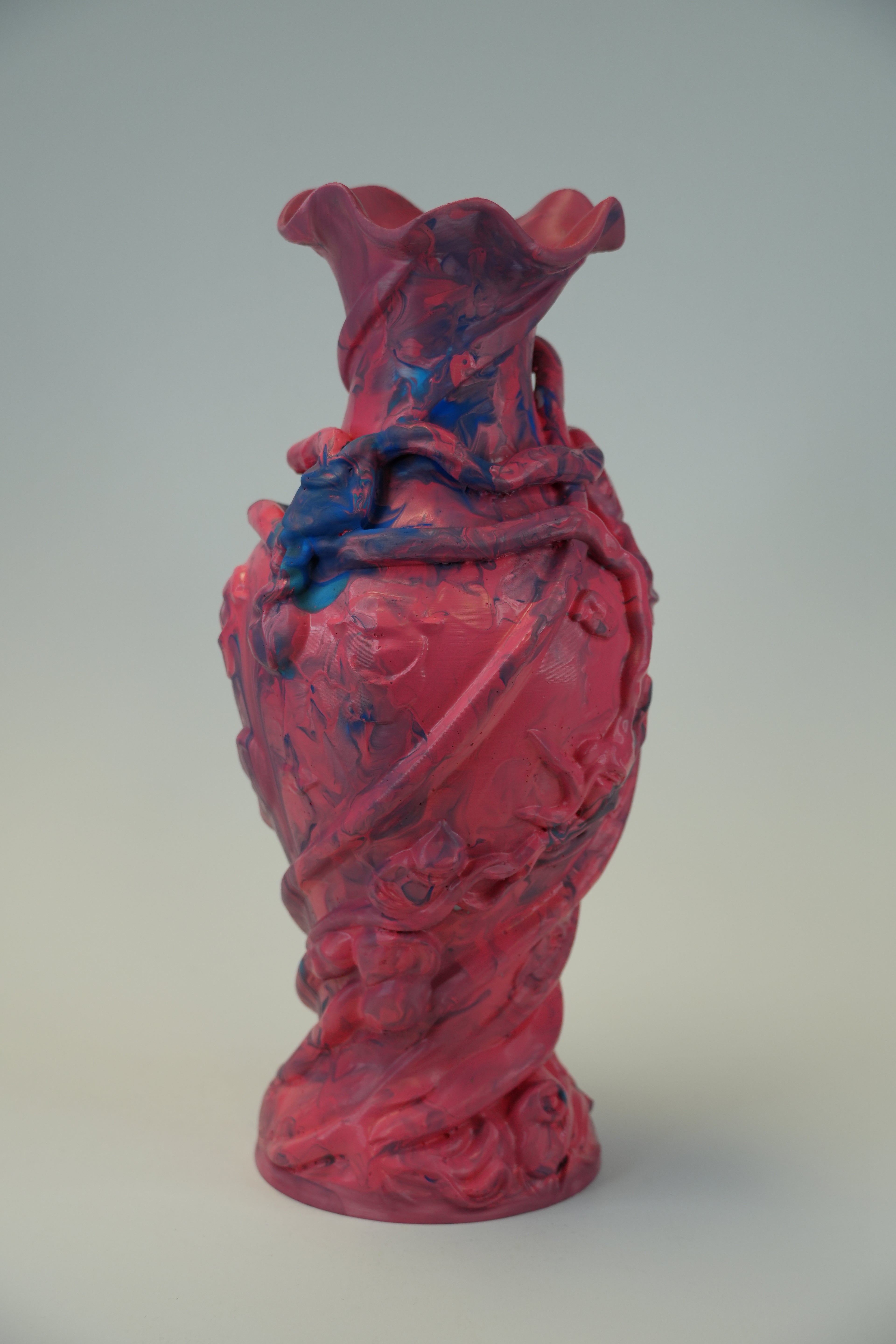 A sculptural ceramic vase with a textured, organic form, featuring a glossy pink and purple body with splashes of blue glaze, set against a neutral gray background.