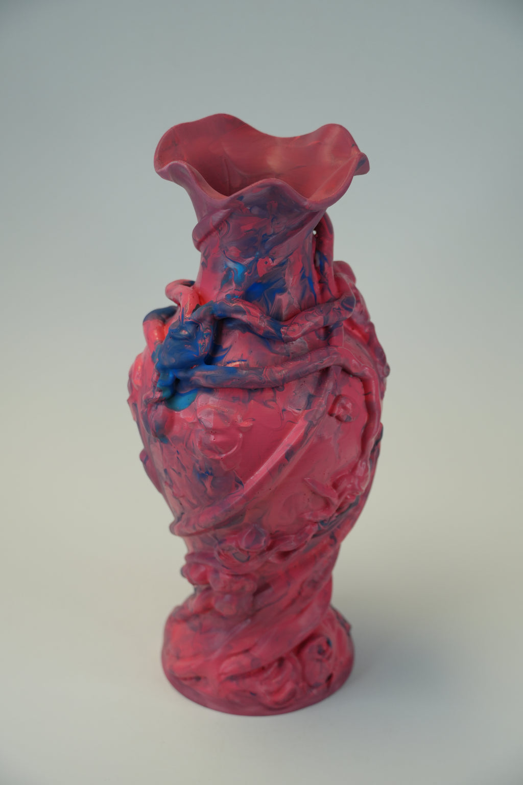 A sculptural ceramic vase with a flared rim and twisted, organic form, painted in a glossy pink-red with splashes of blue and darker purple, photographed against a plain light gray studio background.