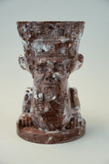 A sculptural brown ceramic planter shaped like a stylized face wearing a crown, photographed against a neutral light gray background.