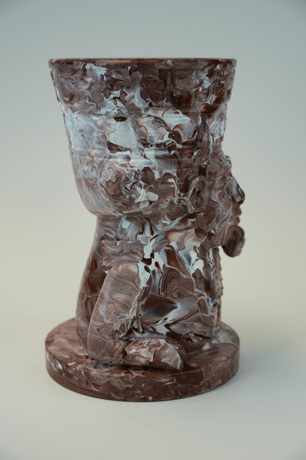 A sculptural, tapered cup or planter made of polished reddish-brown stone with swirling marbled patterns, featuring a carved face profile on the side and a round base, photographed against a neutral light gray background.