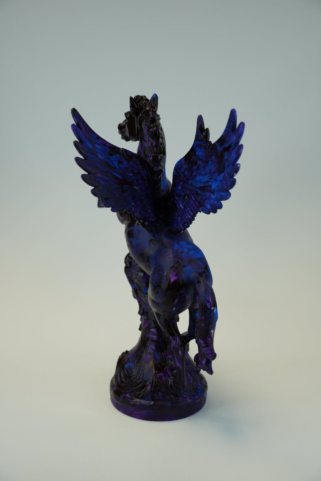A dark blue and purple metallic sculpture of a winged figure, possibly a raven or angel, mounted on a plain light gray wall.