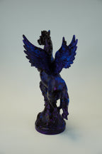 A dark blue and purple metallic sculpture of a winged figure, possibly a raven or angel, mounted on a plain light gray wall.