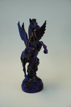 A dark metallic sculpture of a winged horse, likely Pegasus, with a deep blue and black iridescent finish, standing on a matching base against a neutral light gray background.