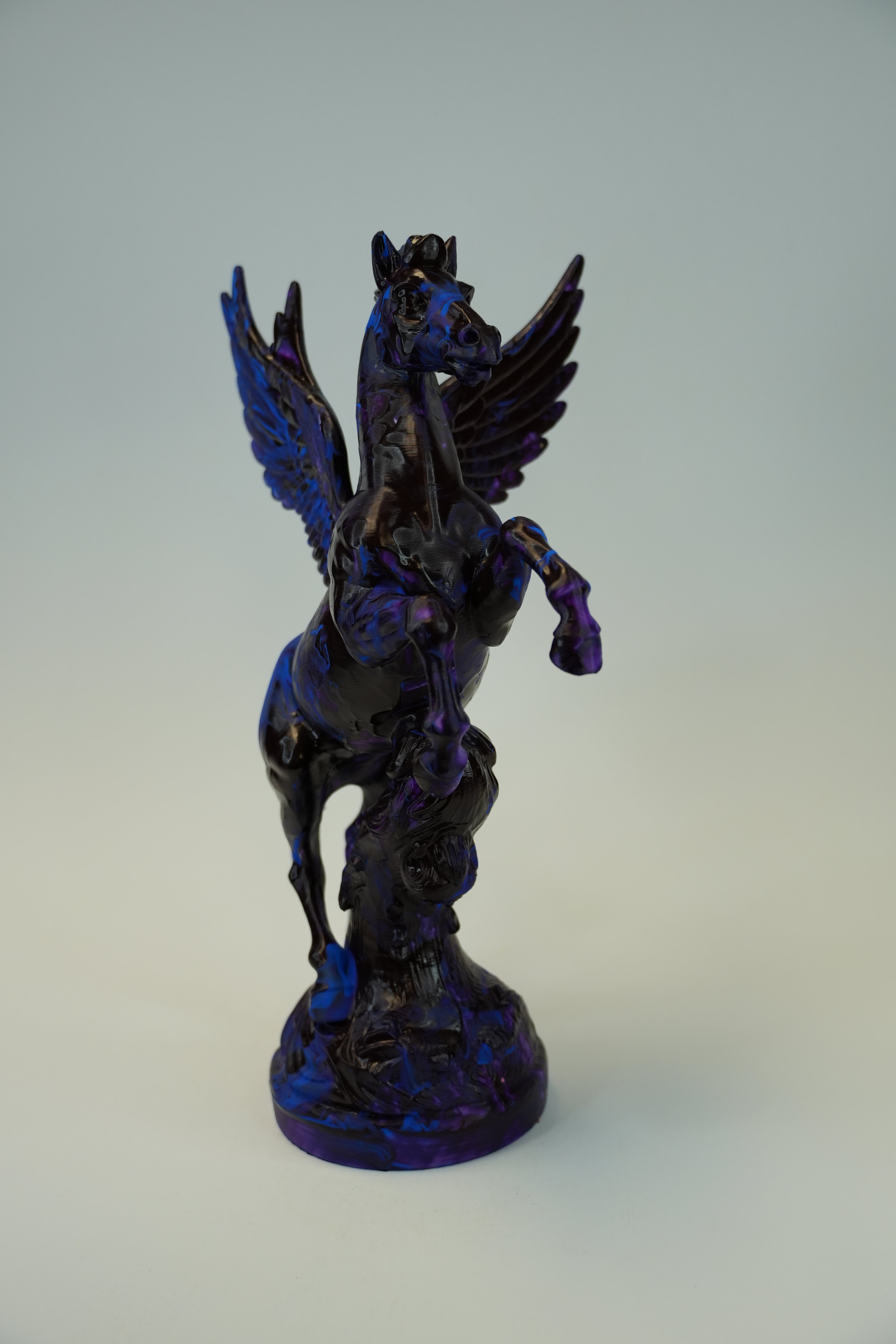 A dark metallic sculpture of a winged horse, likely Pegasus, with a deep blue and black iridescent finish, standing on a matching base against a neutral light gray background.