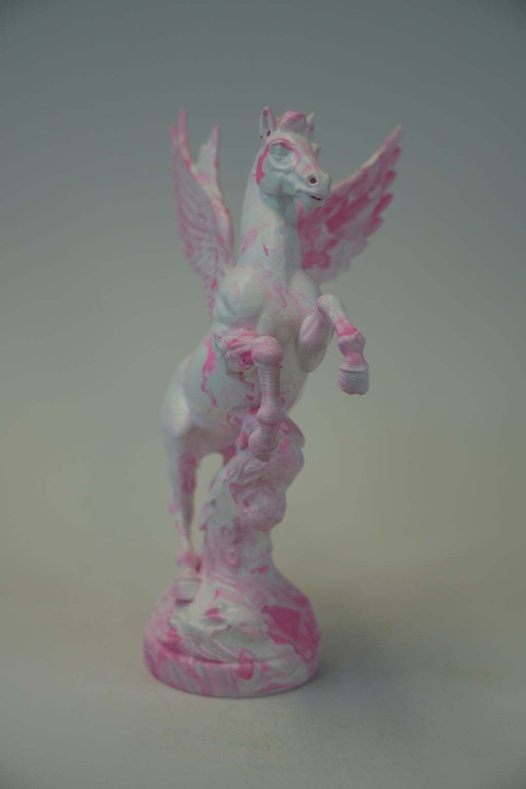 A small white pegasus figurine with pink marbled and splattered details, standing on a matching round base against a plain light gray background.