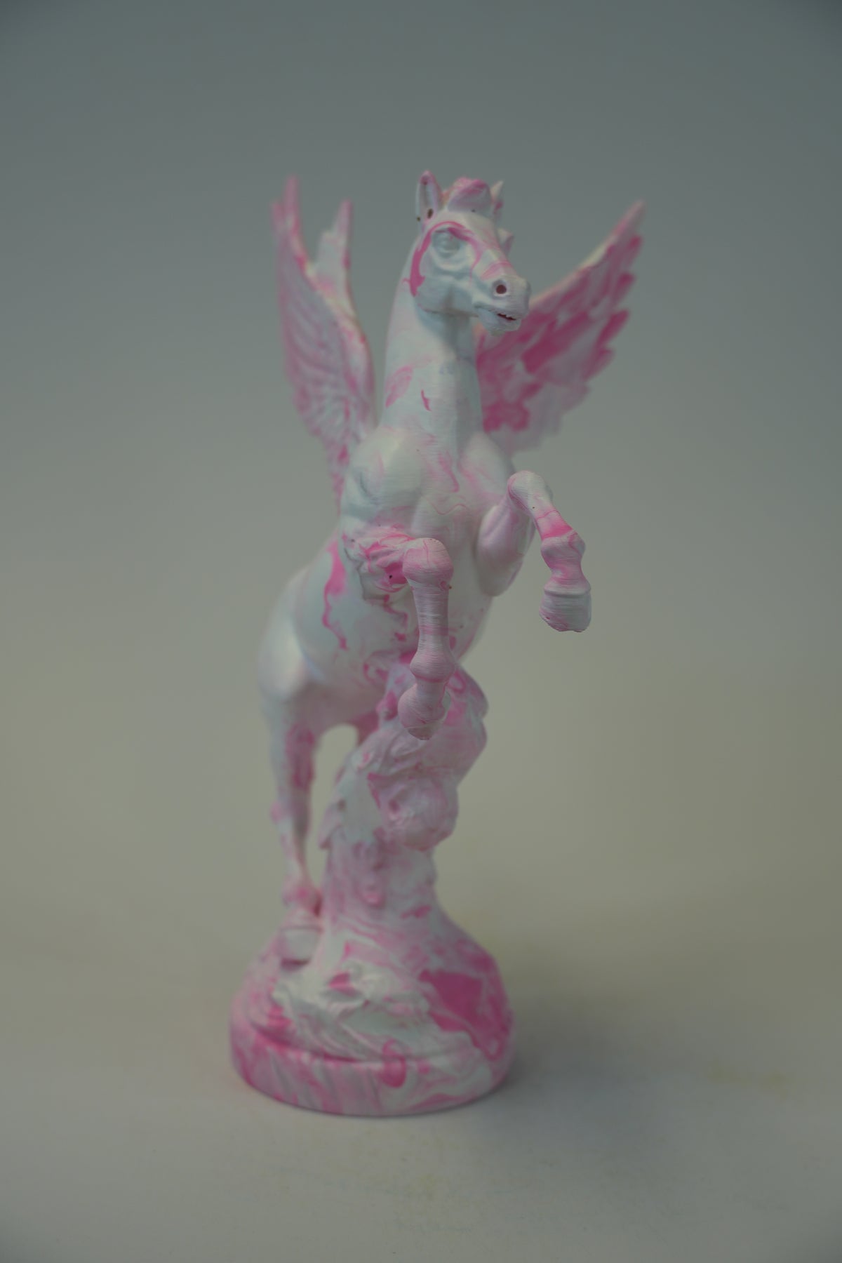 A small white pegasus figurine with pink marbled and splattered details, standing on a matching round base against a plain light gray background.