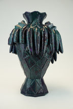 A sculptural ceramic vase with a glossy, iridescent dark green and purple glaze, featuring a textured diamond pattern on the body and a flared, leaf-like rim, set against a plain light background.