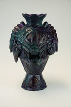 A sculptural ceramic vase with a stylized monster or skull face, featuring a dark iridescent finish that shifts between deep purple, teal, and black, set against a plain light gray background.