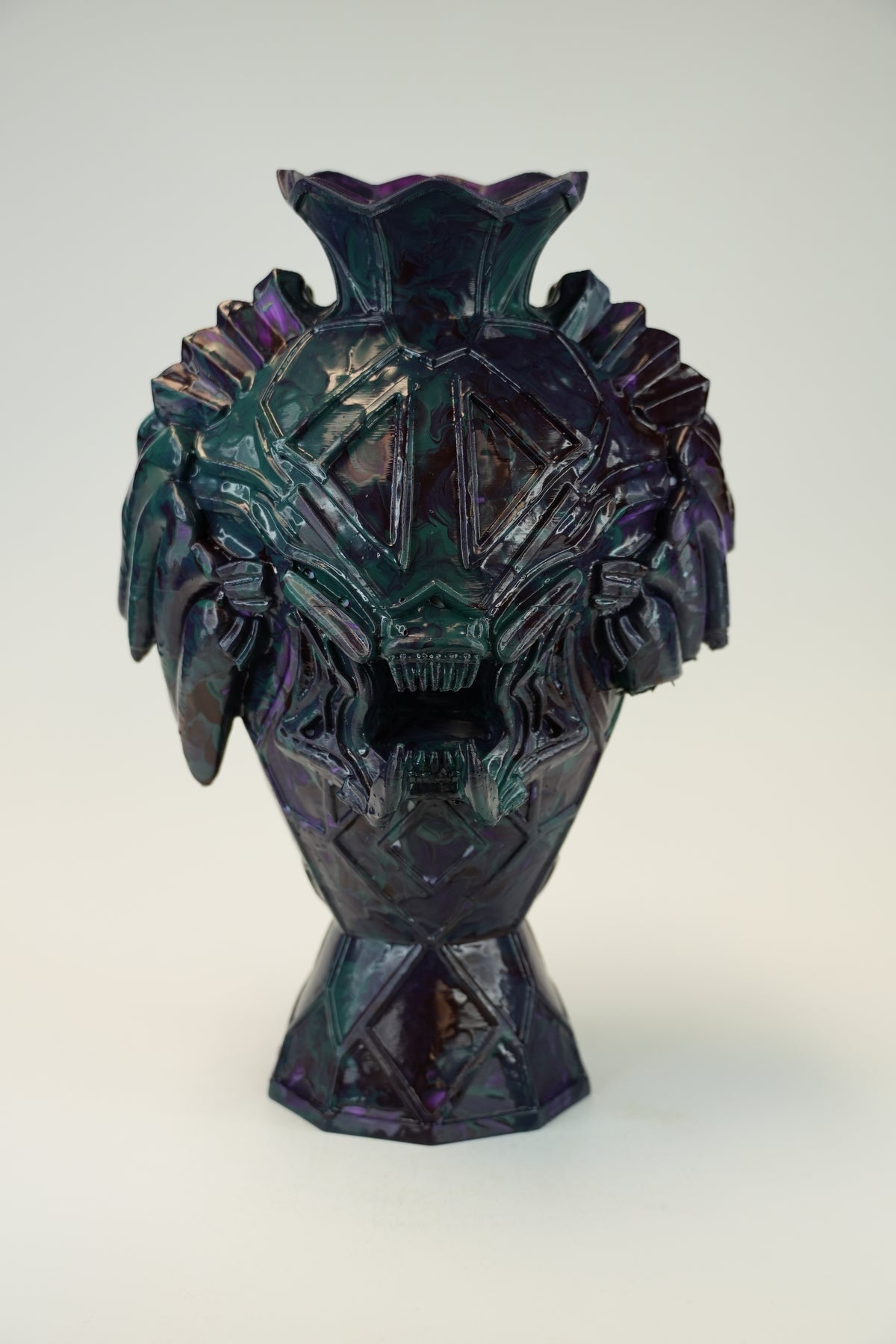 A sculptural ceramic vase with a stylized monster or skull face, featuring a dark iridescent finish that shifts between deep purple, teal, and black, set against a plain light gray background.