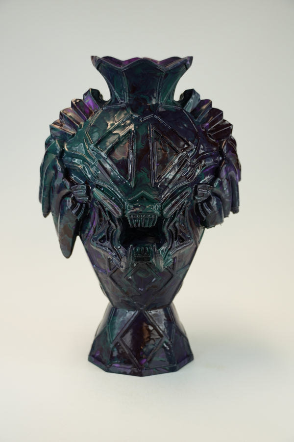 A sculptural ceramic vase with a stylized monster or skull face, featuring a dark iridescent finish that shifts between deep purple, teal, and black, set against a plain light gray background.